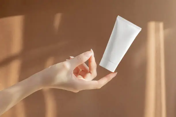 Woman's hand holds white unmarked tube of moisturizer on brown background. Container with anti-aging mask, toothpaste, facial gel. Concept of skin care.