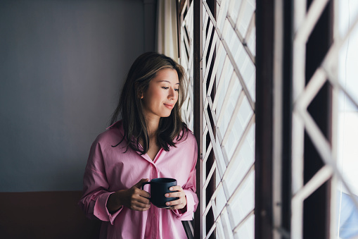 A happy Asian woman drinking her morning coffee and looking out the window.