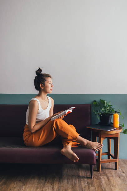 Beautiful Asian Woman in Orange Trousers Sitting on the Sofa and Reading a Book