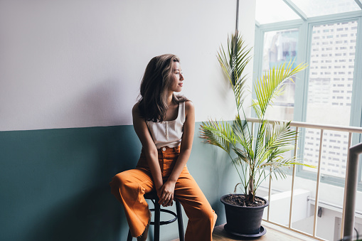 A young Asian woman in orange trousers and a white tank top sitting on a bar chair and looking out the window.