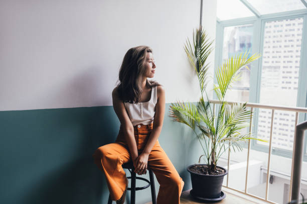 Beautiful Thai Woman Sitting on a Bar Chair and Looking out the Window A young Asian woman in orange trousers and a white tank top sitting on a bar chair and looking out the window. crop top view stock pictures, royalty-free photos & images