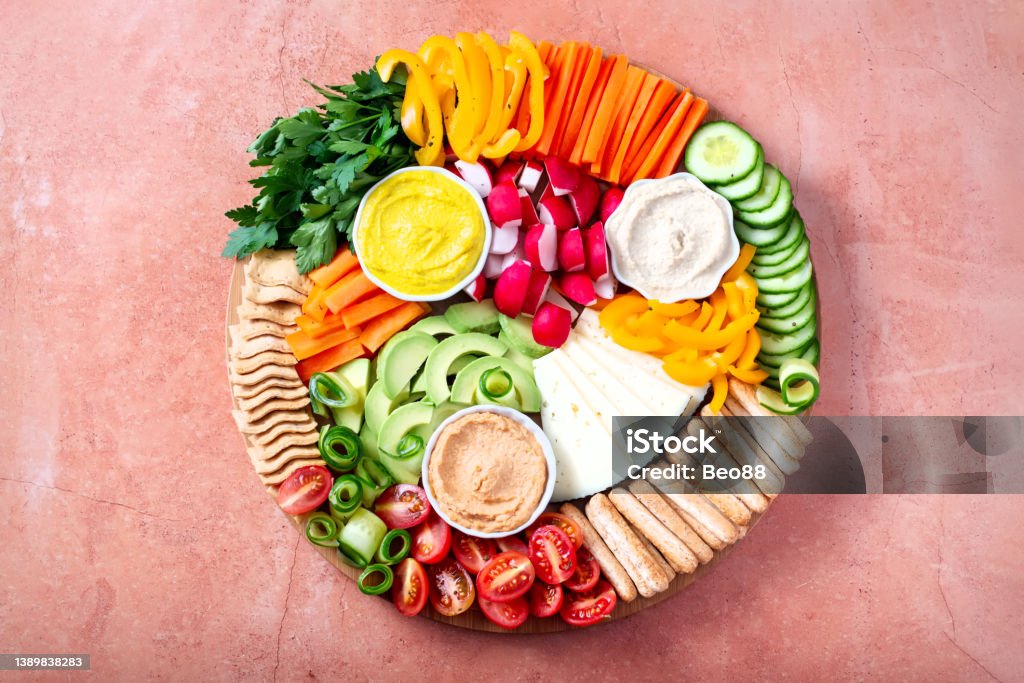 Colorful vegan Charcuterie board with raw vegetables and whole wheat snacks Vegan Charcuterie board with raw vegetables and whole wheat snacks on round board with hummus on stone table, top view, copy space, web banner Plank - Timber Stock Photo