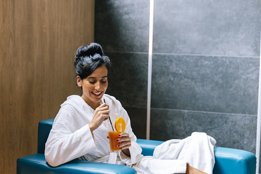 Brunette woman in a bathrobe relaxing in the spa centre, drinking fresh orange juice.