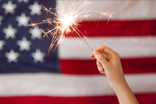 Woman holding bright burning sparkler against American flag, closeup