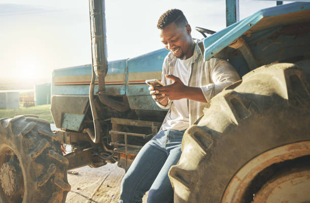 foto de un joven agricultor usando un teléfono mientras está de pie contra un tractor - africa farmer african descent agriculture fotografías e imágenes de stock