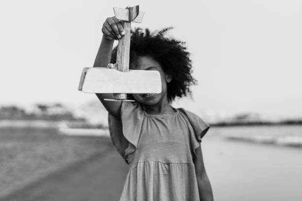 criança africana brincando na praia com avião de madeira - foco na mão segurando brinquedo - edição preto e branco - airplane black and white fun child - fotografias e filmes do acervo