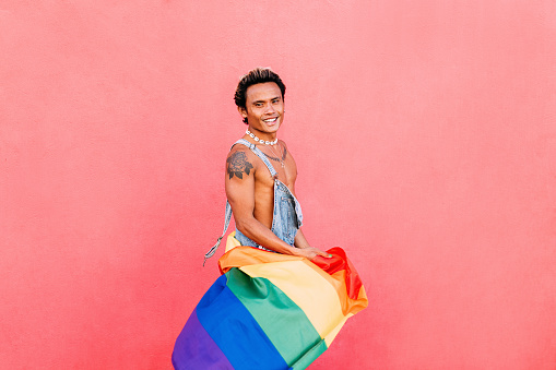 Young handsome guy having fun holding a rainbow flag at wall