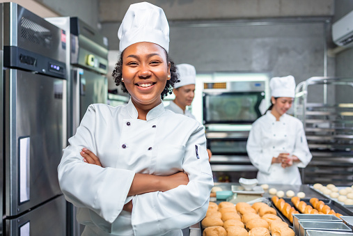 Mujeres panaderas afroamericanas mirando a la cámara. Chef panadero con un vestido de chef y sombrero, cocinando juntos en la cocina. Ella saca galletas recién horneadas del horno eléctrico moderno en la cocina. photo