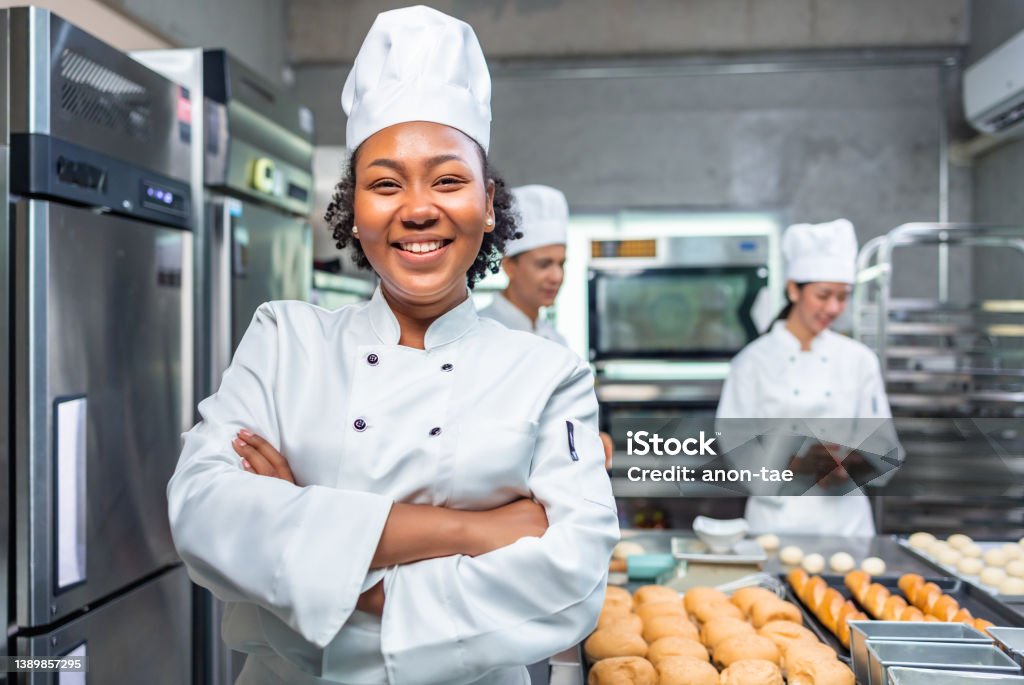 African American woman bakers looking at camera..Chef  baker in a chef dress and hat, cooking together in kitchen.She takes fresh baked cookies out of modern electric oven in kitchen. Chef Stock Photo