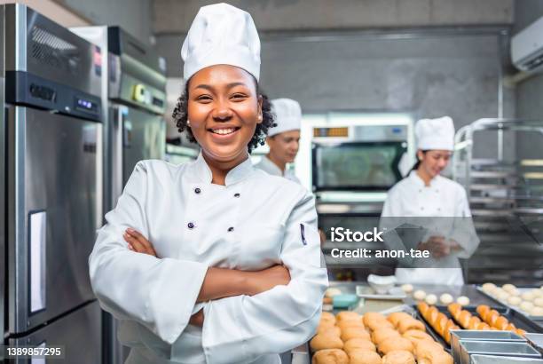 African American Woman Bakers Looking At Camerachef Baker In A Chef Dress And Hat Cooking Together In Kitchenshe Takes Fresh Baked Cookies Out Of Modern Electric Oven In Kitchen Stock Photo - Download Image Now