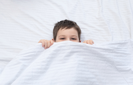 Child boy lies on the bed and looks out from under the blanket. Morning fun at cozy home.