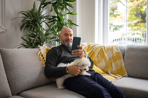 Caucasian senior man using mobile phone while petting Maltese dog