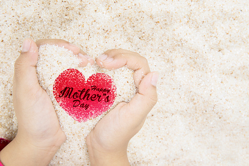 Close up of little girl hands holding sand with happy mothers day text on the heart symbol