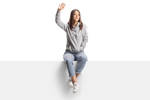 Young female sitting on a blank board and waving at camera isolated on white background
