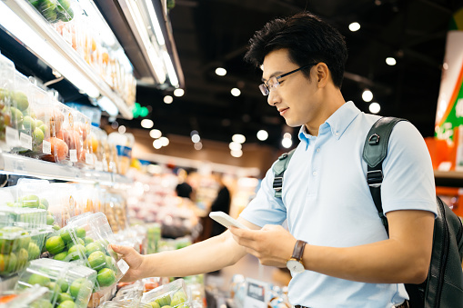 Young Asian man using smart phone  and choosing fresh organic fruits in supermarket.