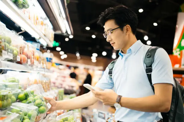 Photo of Young Asian man using smart phone  and choosing fresh organic fruits in supermarket.
