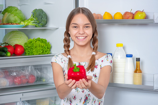 Teen girl holding fresh red pepper while standing near open fridge in kitchen at home