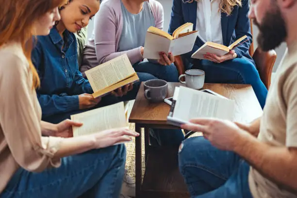 Small group of people with a mixed age range sitting at a table, discussing and reading books together.