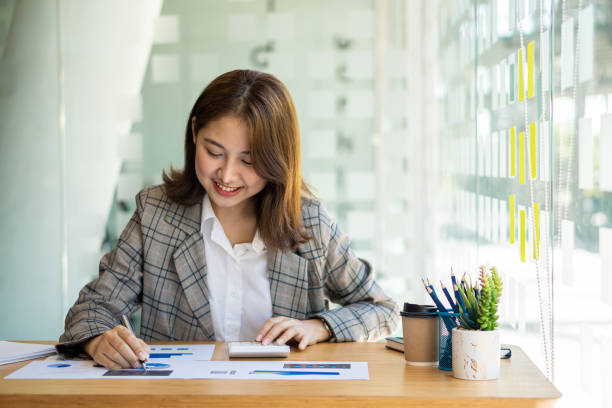 istock Beautiful Asian businesswoman analyzing chart using calculator, laptop at the office with papers and holding pen, sitting on the chair at modern home studio. Mobile worker contacting customers 1389877047