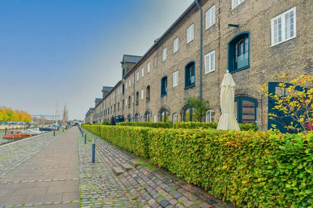 Many three-story buildings made of gray bricks on a street with a stone pavement near the river channel on the neighbourhood Christianshavn. Copenhagen, Denmark
