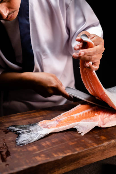 mano dello chef che tiene un pezzo di salmone fresco. primo piano delle mani dello chef che preparano cibo giapponese. chef giapponese che prepara sushi al ristorante. chef che prepara sushi tradizionale giapponese su tavola di legno. - thai cuisine asian cuisine vertical close up foto e immagini stock