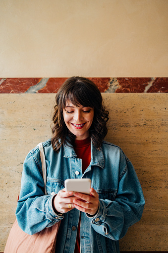 Happy woman with dental braces typing text message on her smartphone while standing in front of the wall.