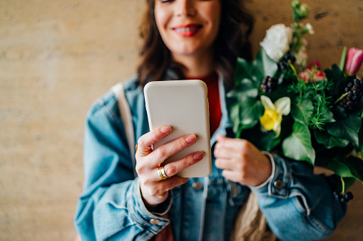 Close up photo of woman hands holding bouquet of flowers and typing text message on her smartphone while standing in front of the wall.