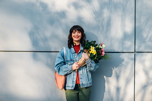 Smiling woman with dental braces looking at camera while holding bouquet of flowers and standing  in front of the gray wall.