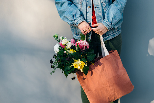 Close up photo of woman hands holding bouquet of flowers in her bag while standing in front of the gray wall.