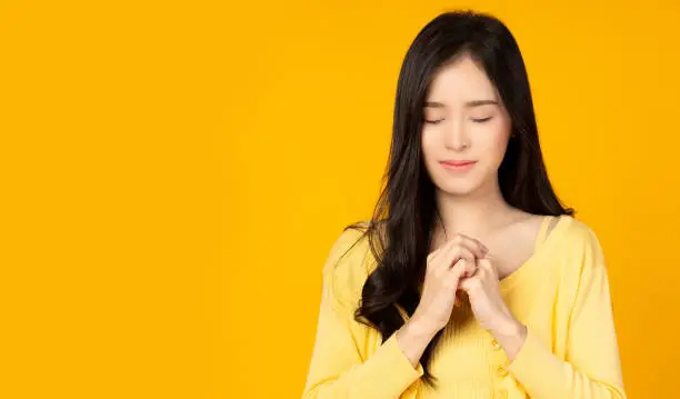 Photo of Young asian millennial woman hold hands on heart chest Beautiful girl get grateful and thankful Happy lady show gratitude, love and care, pray or make a wish Religion and faith concept She closed eyes