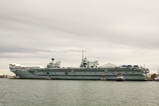 The Queen Elizabeth aircraft carrier moored at Navy dockyard in Portsmouth, Hampshire, England.. With tugs attending. View from boat on the water.