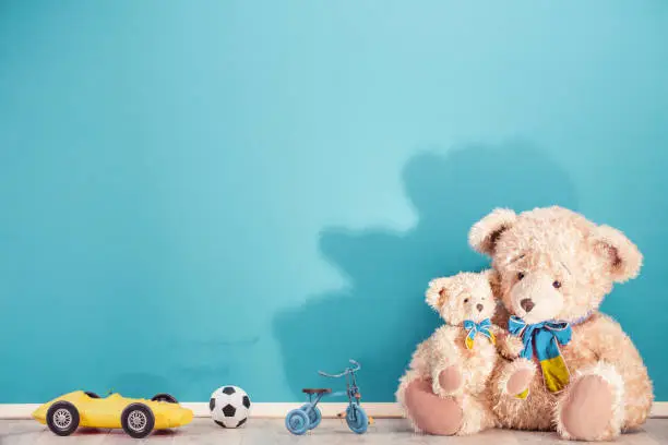 Photo of Teddy Bears family, old toy trike bicycle, obsolete retro yellow plastic car, soccer ball front blue wall with shadow background. Mother or father with baby concept. Vintage style filtered photo