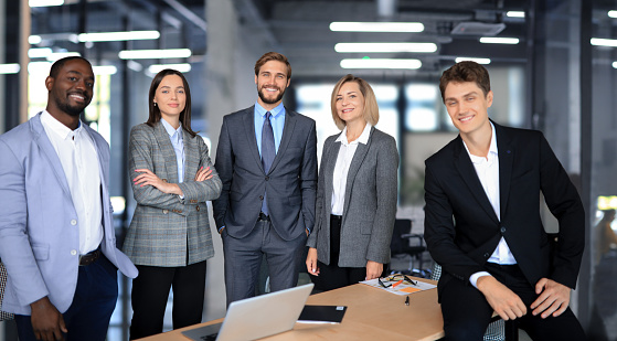 Team of business people posing for a photo. They are all smiling and looking at the camera. They are standing and wearing formal business clothing. There are several ethnicities in the group.