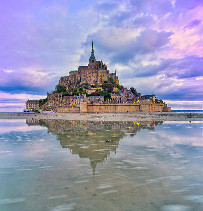 Normandy, France, August 29, 2019: Reflection on the out-flowing water, during low tide give a special perspective of the famous Mont Saint-Michel castle. Le Mont-Saint-Michel or in english Saint Michael's Mount is a tidal island and mainland commune in Normandy, France. The island lies approximately one kilometre (0.6 miles) off the country's north-western coast, at the mouth of the Couesnon River near Avranches and is 7 hectares (17 acres) in area. The mainland part of the commune is 393 hectares (971 acres) in area so that the total surface of the commune is 400 hectares (988 acres). As of 2017, the island has a population of 30. \nAs of 2017, the island has a population of 30. Mont-Saint-Michel and its bay are on the UNESCO list of World Heritage Sites. It is visited by more than 3 million people each year. Over 60 buildings within the commune are protected in France as monuments historiques.