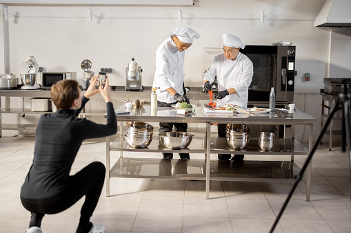 Man records on phone the process of cooking by two chefs in a professional kitchen. Latin-American and Asian cooks preparing food for social media. Concept of culinary video blogging