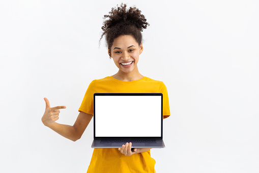 Portrait of young African American woman with laptop on white background. Beautiful female points her finger at a blank laptop screen, looking at the camera and smiling