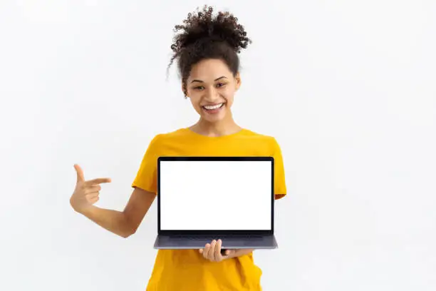 Photo of Portrait of young African American woman with laptop on white background. Beautiful female points her finger at a blank laptop screen, looking at the camera and smiling