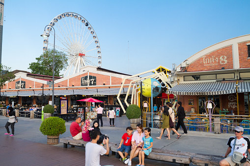 Late afternoon scene on promenade at Asiatique at Chao Praya river in Bangkok. People are walking between restaurants. Behind buildings is a ferris wheel. In foreground a man is photographing family. Asiatique is promenade area with restaurants in southern Bangkok