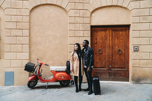 Young adult couple is standing in a street of Siena near an old scooter, waiting for a taxi. They are looking away.