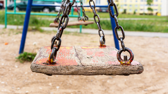 old wooden swing on rusty metal chains in local playground against blurry city buildings and parking lot close view