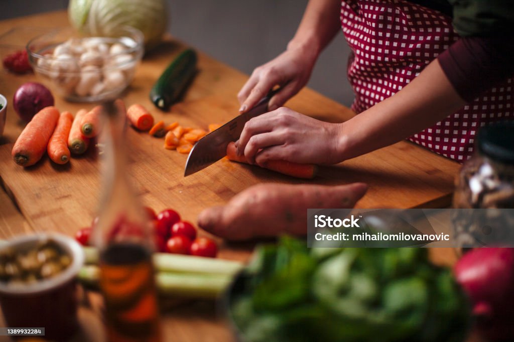 Slicing carrots Hands of young woman as she chopping carrots with big knife with various vegetables around on kitchen counter Vegetable Stock Photo