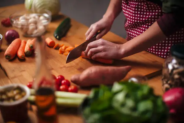 Hands of young woman as she chopping carrots with big knife with various vegetables around on kitchen counter