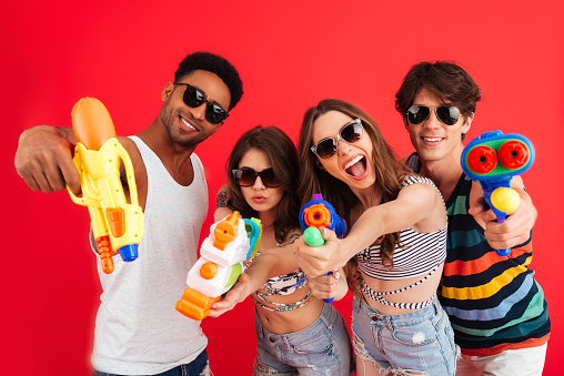 Portrait of a happy smiling multiracial group of friends in summer clothes posing while standing with water pistol isolated over red background