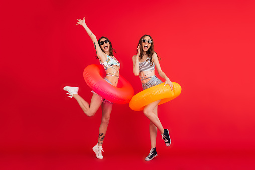 Full length portrait of two excited smiling girls in sunglasses jumping with inflatable rings isolated over red background