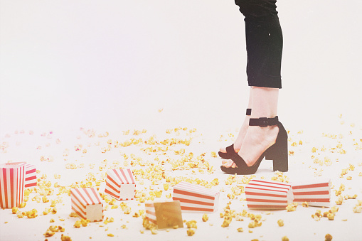 Leisure and lifestyle concept. Many popcorn and boxes tossed on floor and woman legs with black trousers and high heel shoes in white background. Vintage and grunge filters with grain, noise applied