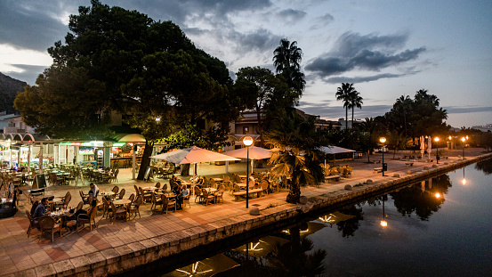 Alcudia, Majorca, Balearic Islands, Spain – October 16, 2018: Restaurants, cafes, and hotel complex outdoors near the river at midnight with evening lights. Palms, sky and people around. European landscape and lifestyle photography