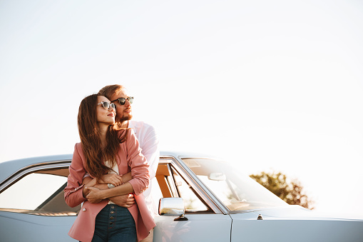 Young smiling couple hugging while standing near the retro car and looking away
