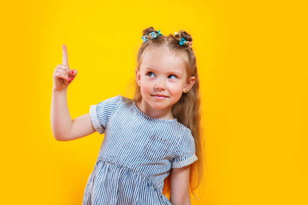 istock Little girl is looking pointing forefingers up tips feedback good solution isolated over yellow background 1389960254