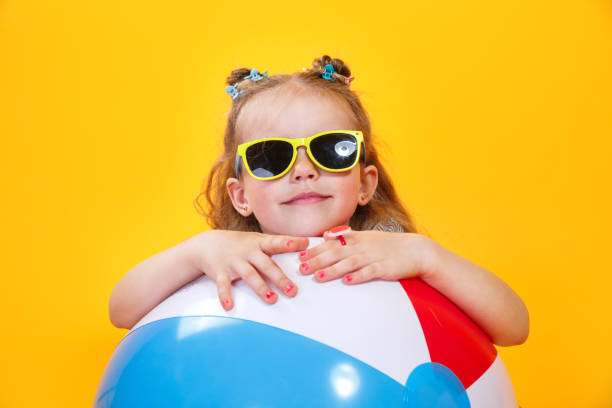 Summer girl wearing sunglasses with ball laughing on yellow background stock photo