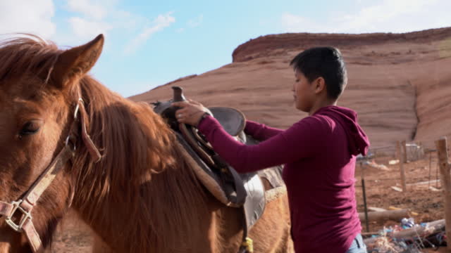 Pretty Teenage Navajo Girl with Short Hair Saddles Horse with Saddle Blanket near Navajo Tribal Park in Monument Valley Arizona